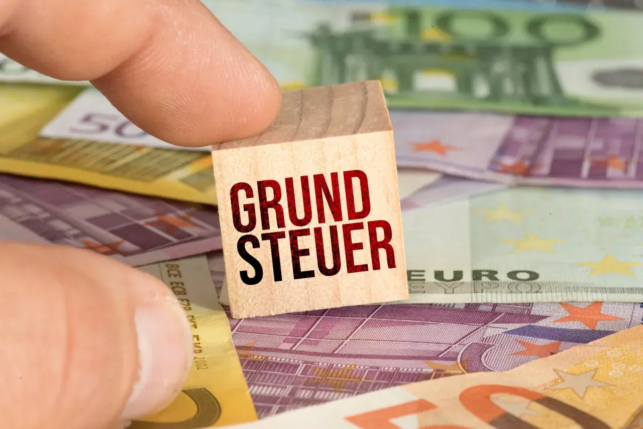 Ein Finger hält einen Holzstein mit der Aufschrift "GRUNDSTEUER" über Euro-Banknoten. Der **Grundsteuer Messbetrag** ist hier visuell dargestellt.