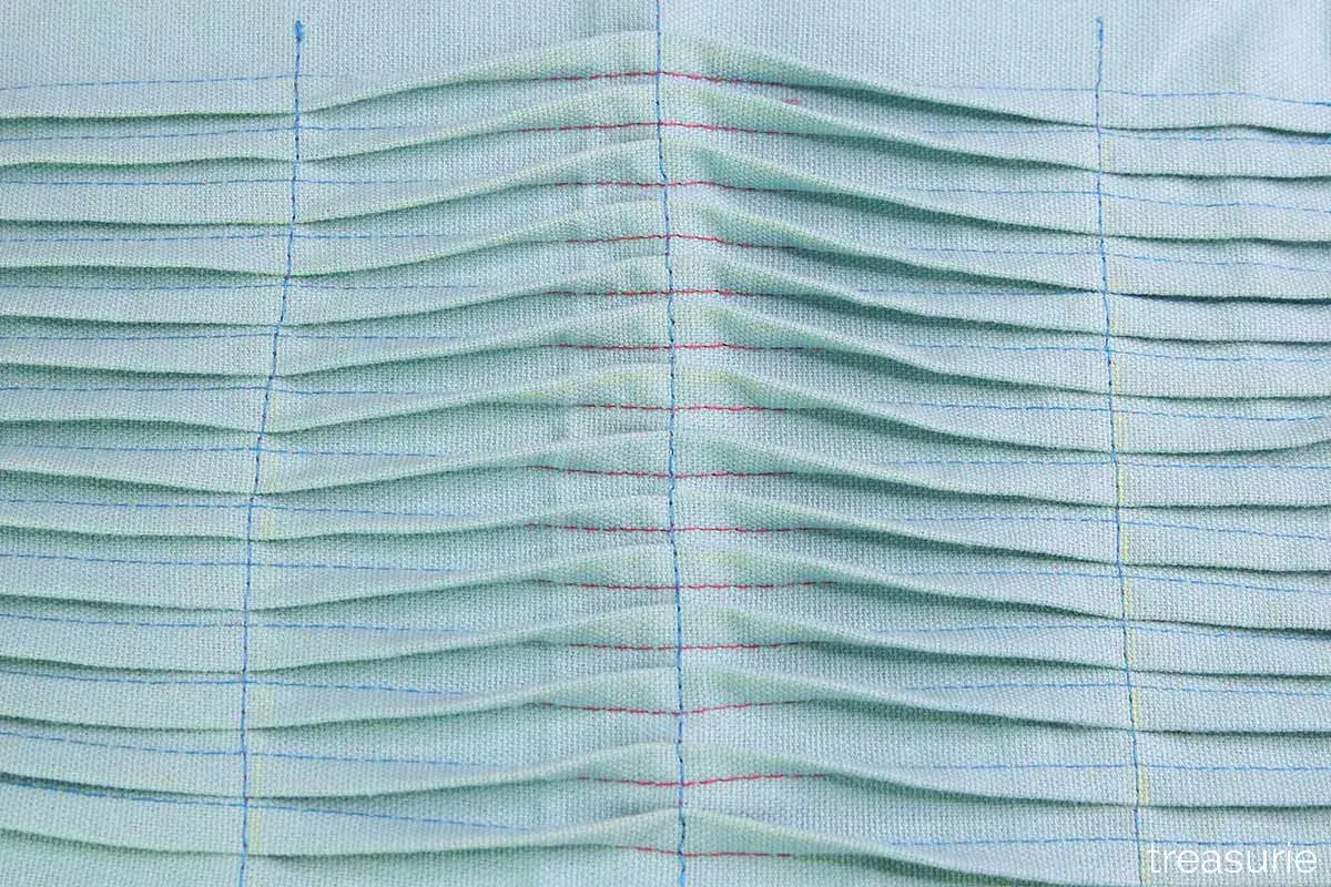 Close-up of light blue fabric with rows of pintucks, creating a textured, pleated effect. Red and blue threads highlight the stitching.