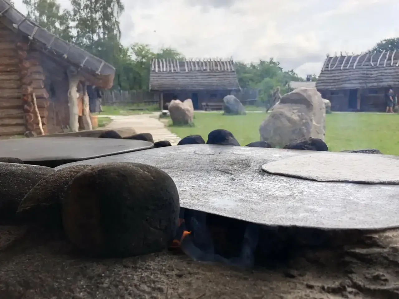 Ognisko pod płaskim kamieniem, na którym piecze się podpłomyk. W tle drewniane chaty i zielony dziedziniec osady średniowiecznej Sławutowo.
