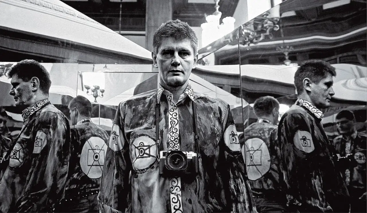 Krzysztof Miller w lustrzanym odbiciu, z aparatem na szyi, w koszuli z motywem oka i aparatu.