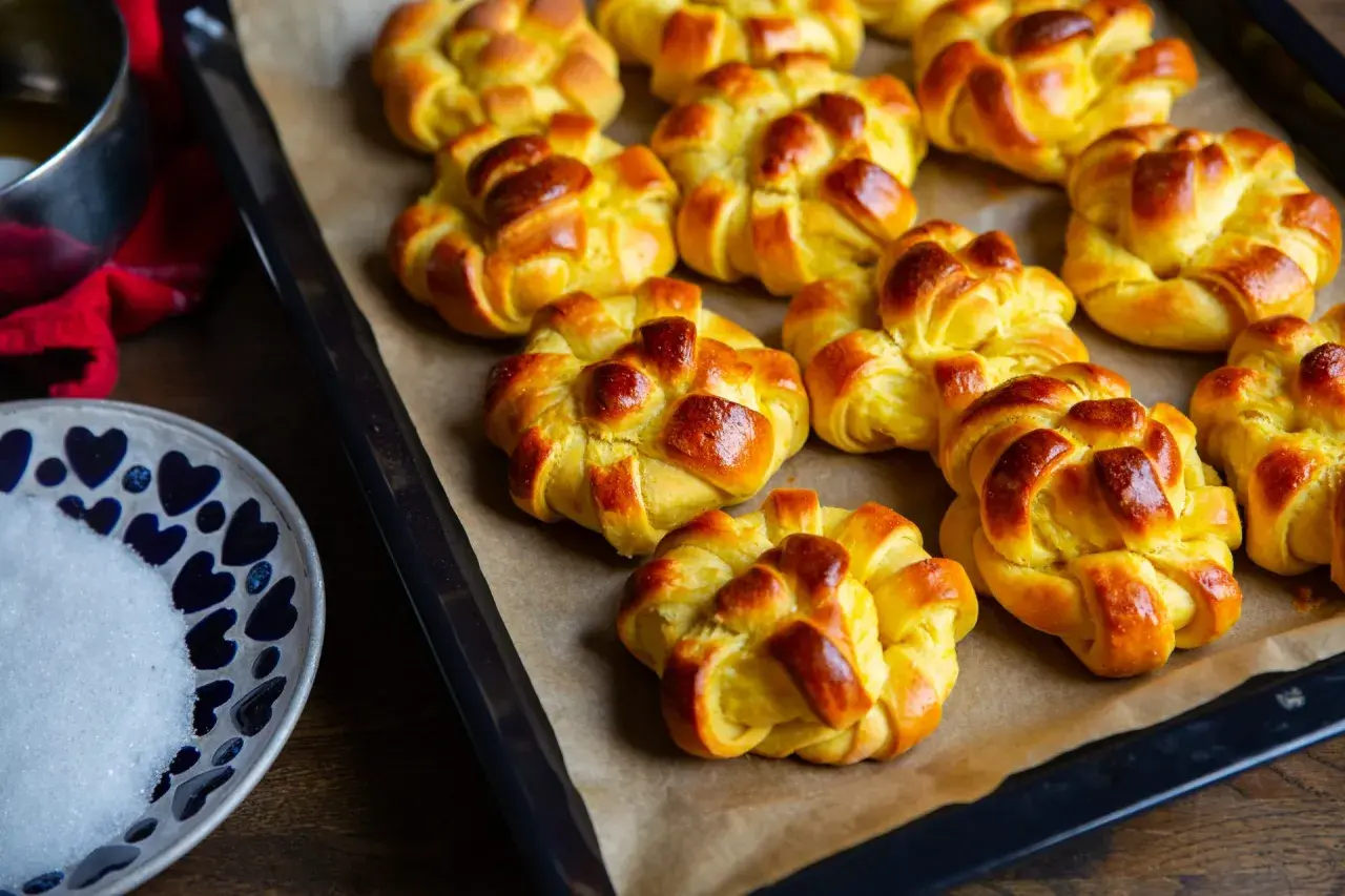 Nybakade zeina saffransbullar på en plåt, gyllene och inbjudande. En skål med socker och en röd duk anas i bakgrunden.