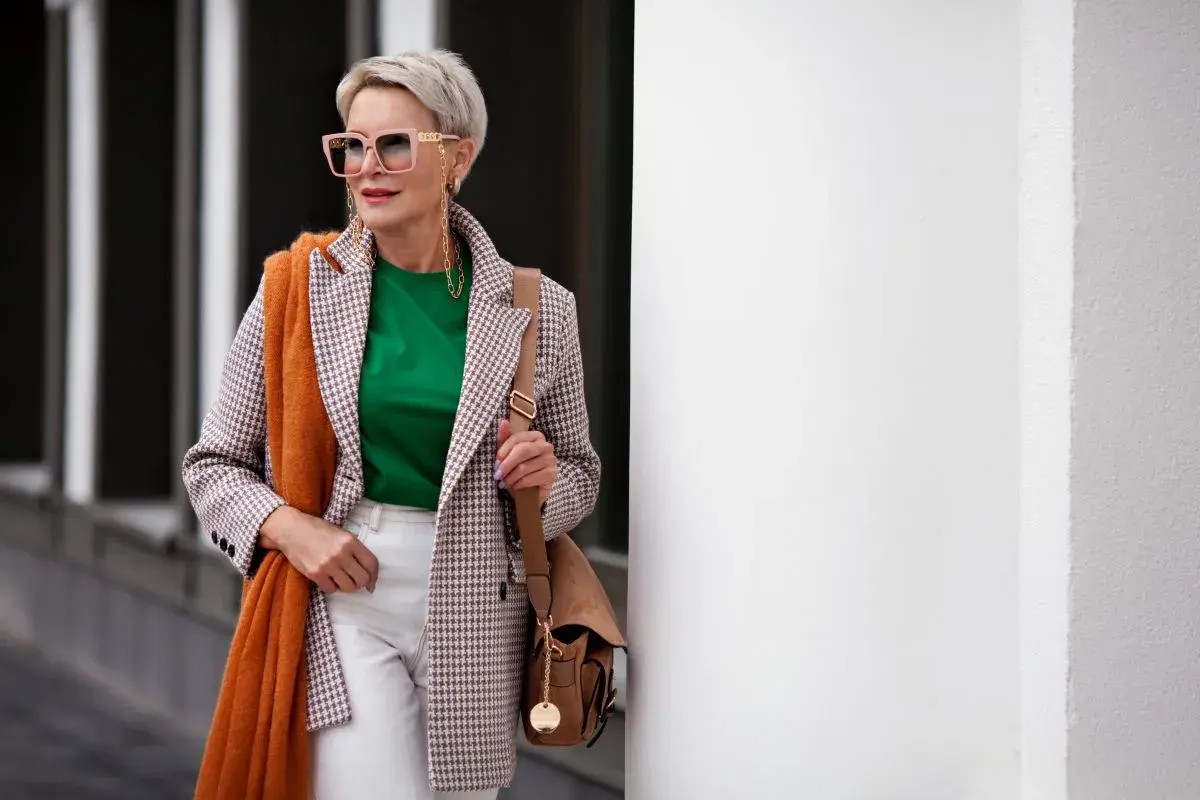 Elegancka kobieta po 60. w stylowej marynarce w kratkę, zielonej bluzce i białych spodniach. Pomarańczowy szal dodaje jej uroku.