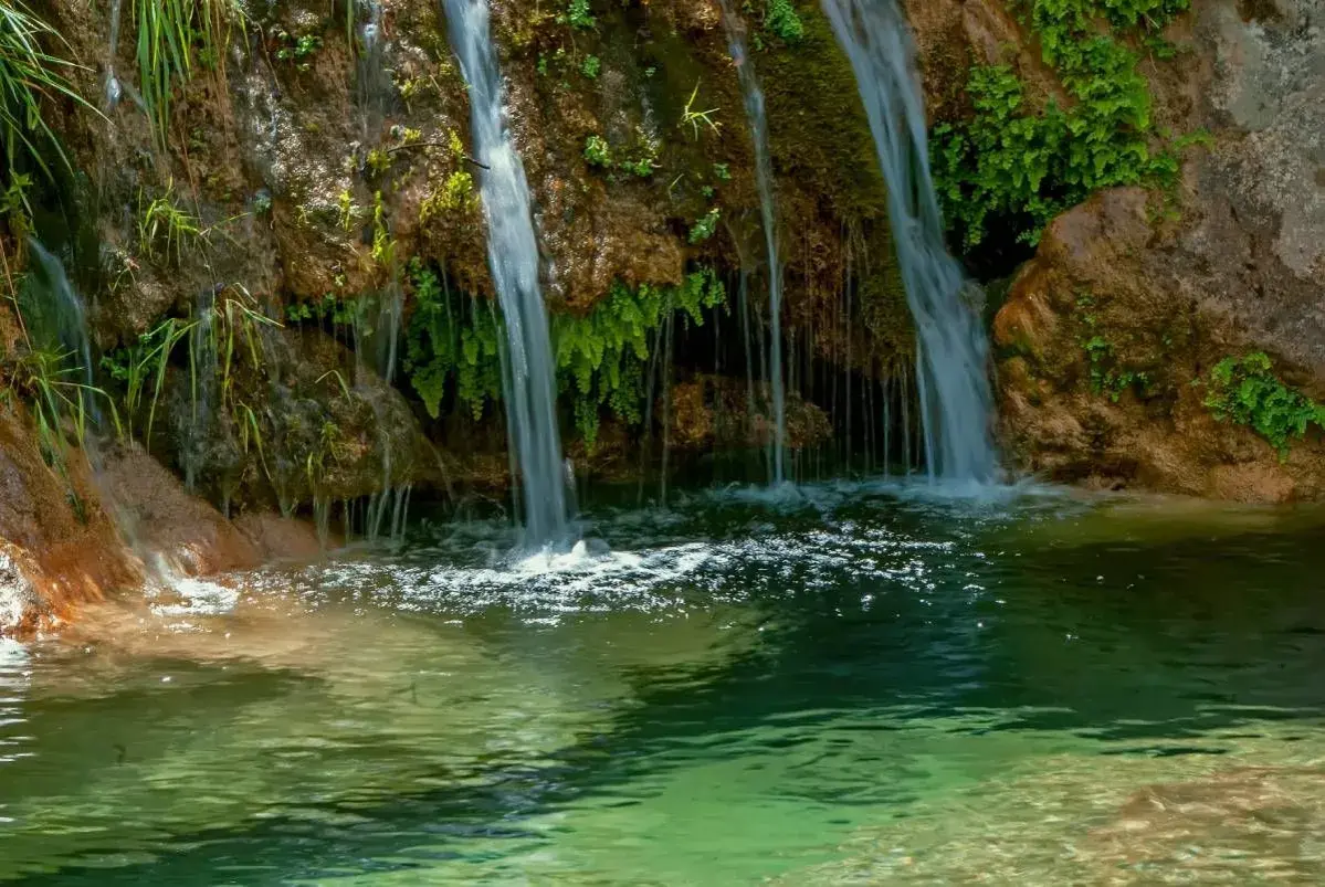 Cascada de la Osera, con agua cayendo sobre rocas cubiertas de musgo y vegetación exuberante, formando un estanque de aguas cristalinas.