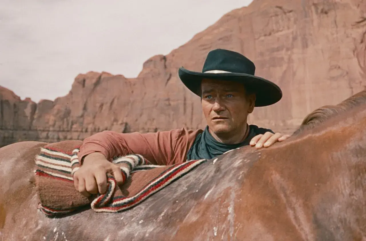 John Wayne w kapeluszu, oparty o konia, w krajobrazie przypominającym miasto bezprawia.