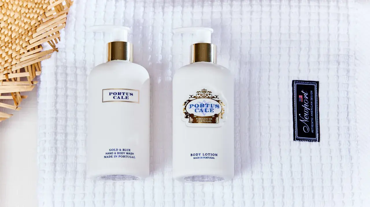 Two white pump bottles of Portus Cale body wash and lotion on a white textured towel, with a woven straw fan and a Newport tag.