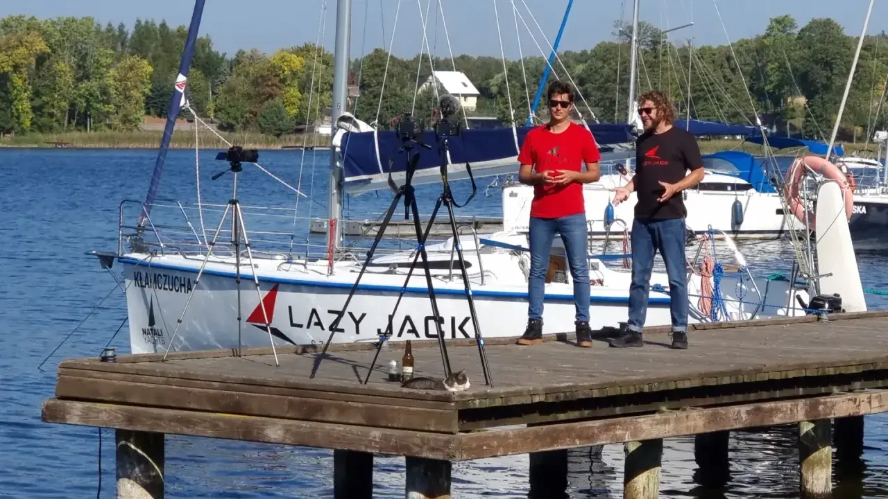 Dwóch mężczyzn na pomoście nagrywa film na tle żaglówki "Lazy Jack". Na pomoście leży kot.