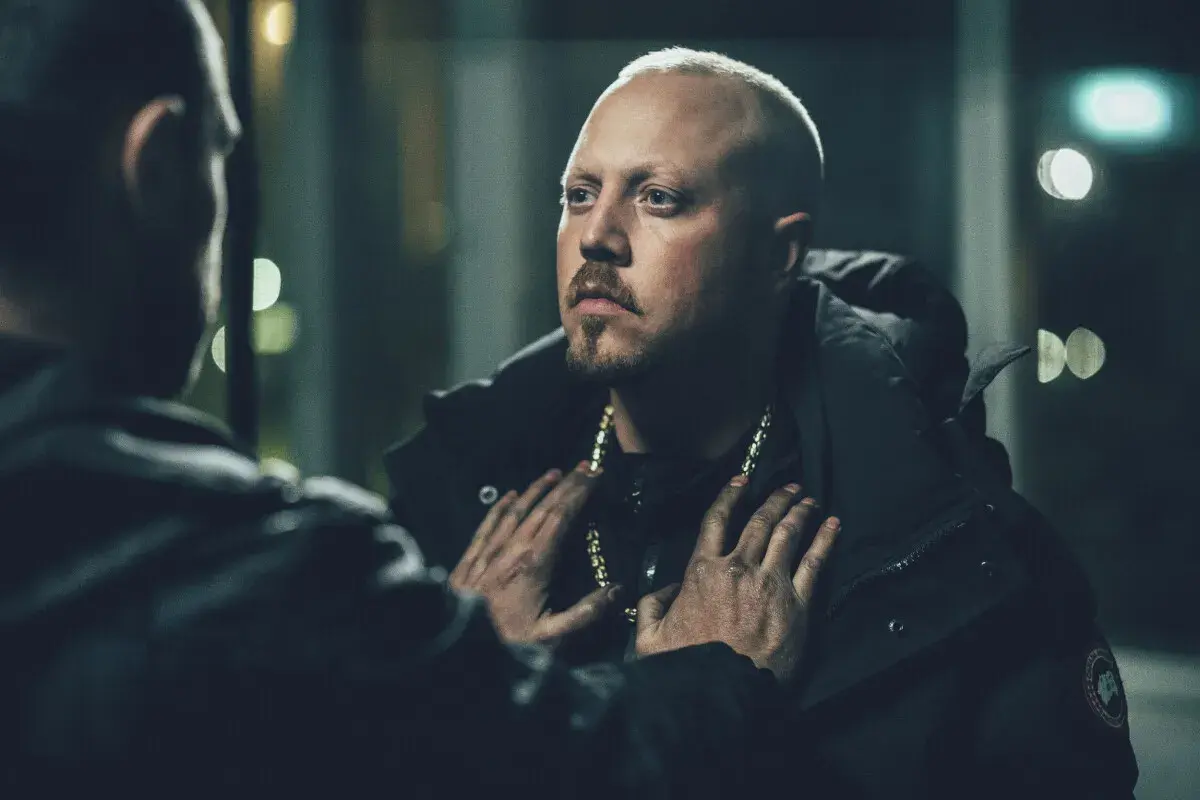 En dramatisk scen från en sebbe stakset film. Två män möts i mörkret, en med guldhalsband, den andra med händerna på hans bröst.