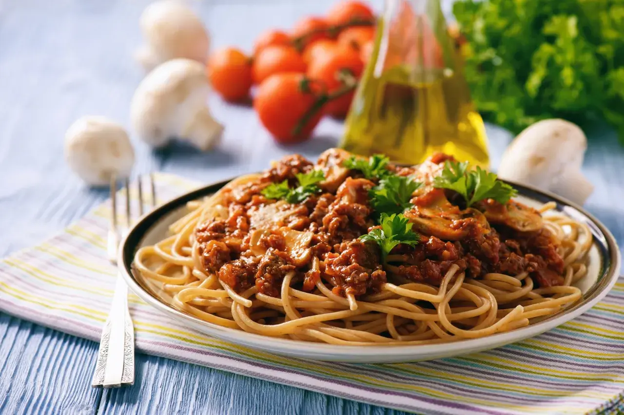 Une assiette de tagliatelles à la bolognaise, garnie de persil frais. Des champignons, tomates et une bouteille d'huile d'olive sont en arrière-plan. Une recette parfaite pour un repas réconfortant.