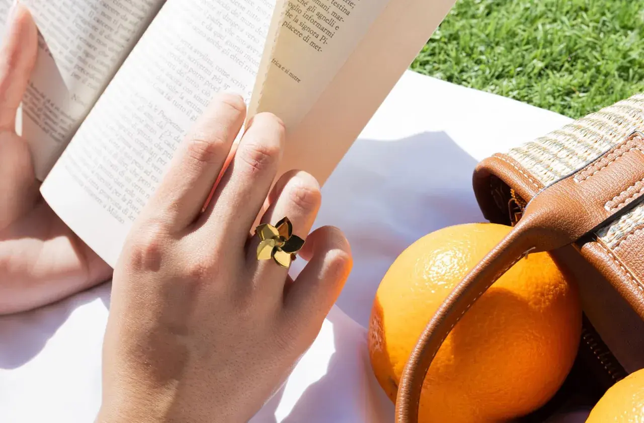 Mano con un anillo dorado con forma de flor, leyendo un libro junto a naranjas y un bolso de mimbre. Las tallas de anillos perfectas para cada ocasión.