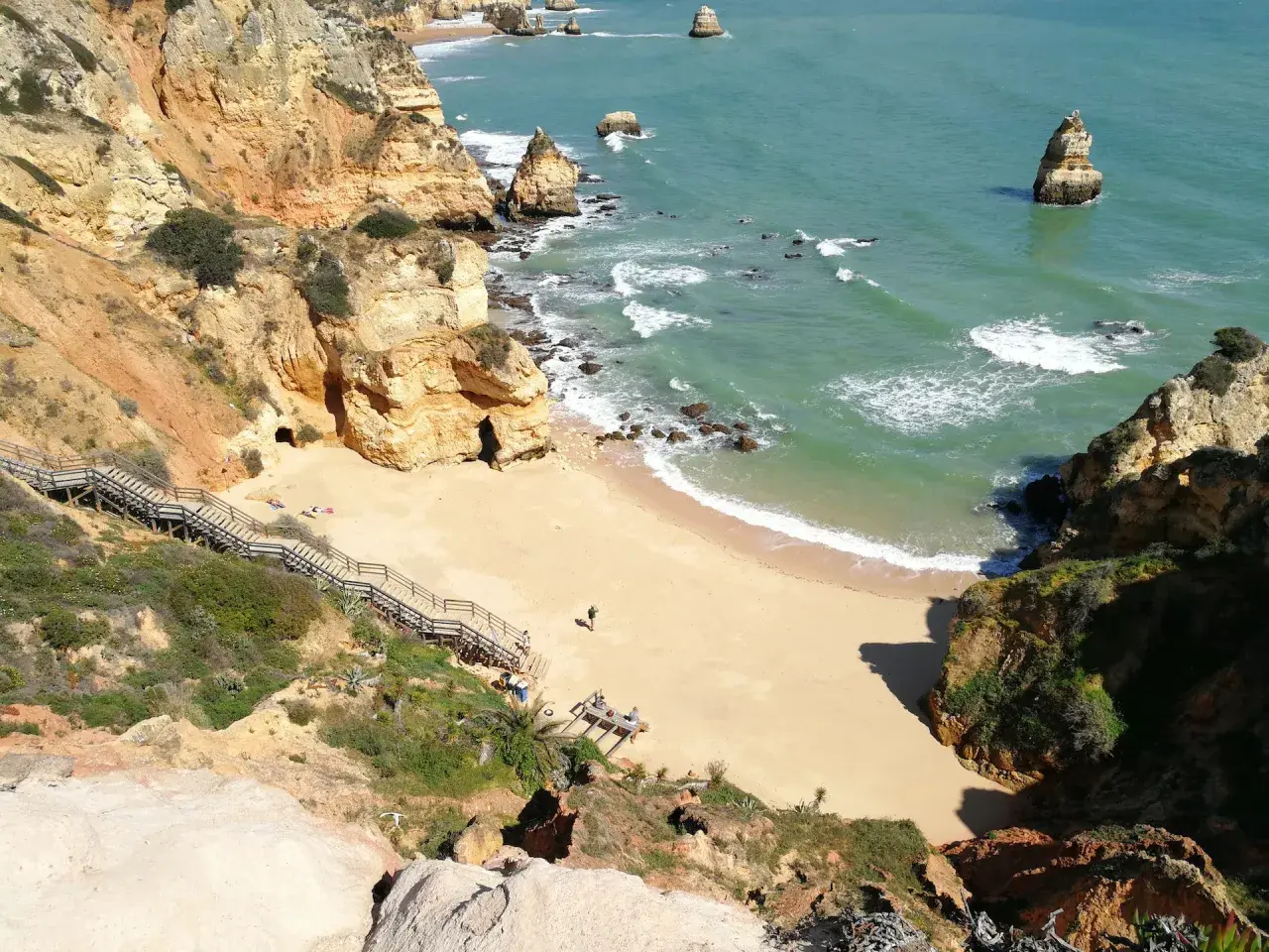 Acantilados dorados y mar turquesa en las **islas de Portugal**. Escaleras de madera descienden a una playa de arena dorada, con formaciones rocosas emergiendo del agua.