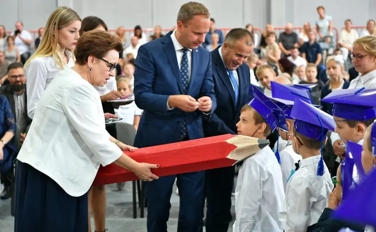 Kobieta wręcza dzieciom w biretach gigantyczny czerwony ołówek.