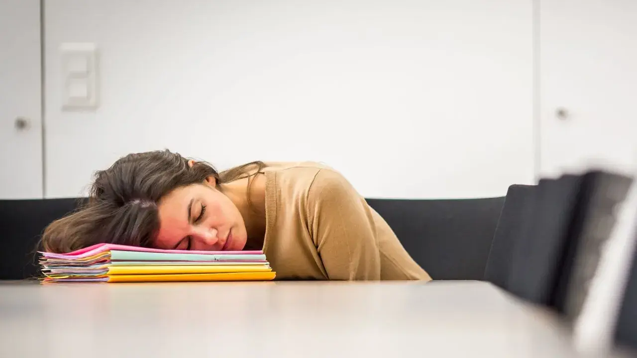 En kvinna sover uttråkad på jobbet, med huvudet vilande på en hög med färgglada mappar.