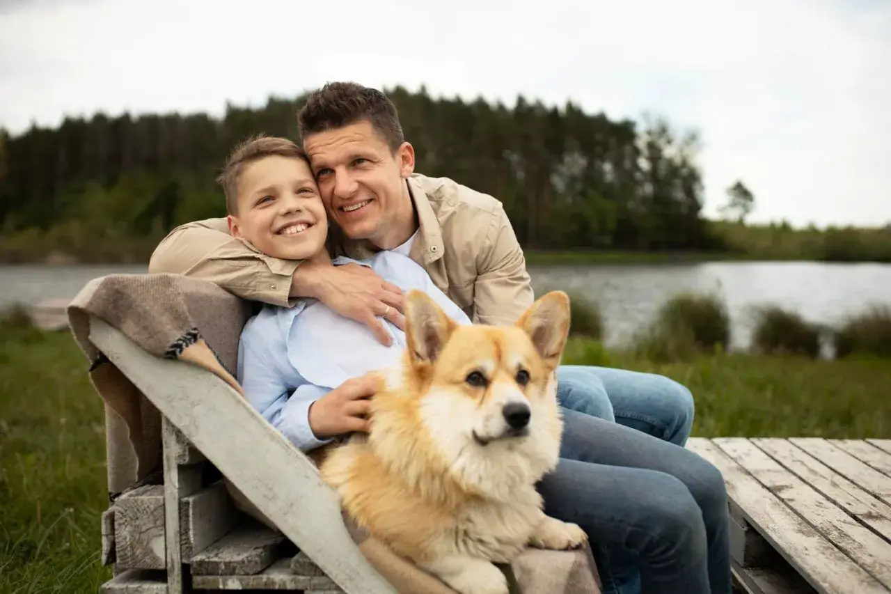 Ojciec i syn siedzą przy jeziorze z psem. Corgi kładzie się na kolanach chłopca, bo czuje się bezpiecznie i kochany.