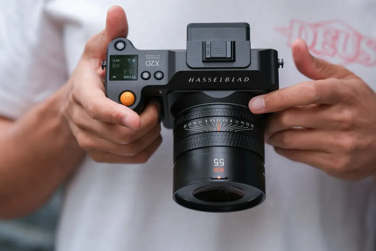 Dłonie trzymają aparat Hasselblad X2D 100C z obiektywem XCD 55mm. Widoczne detale aparatu i obiektywu.