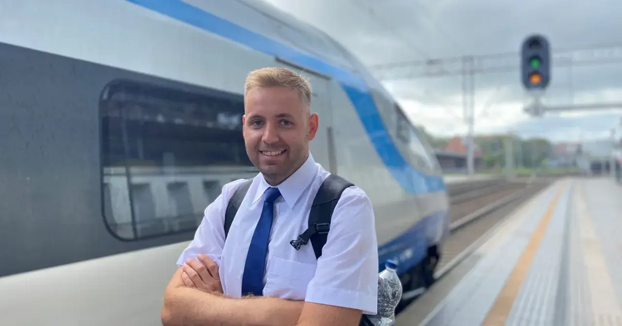 Uśmiechnięty mężczyzna w białej koszuli i niebieskim krawacie, gotowy do pracy. To on prowadzi pociąg, który stoi obok na torach.