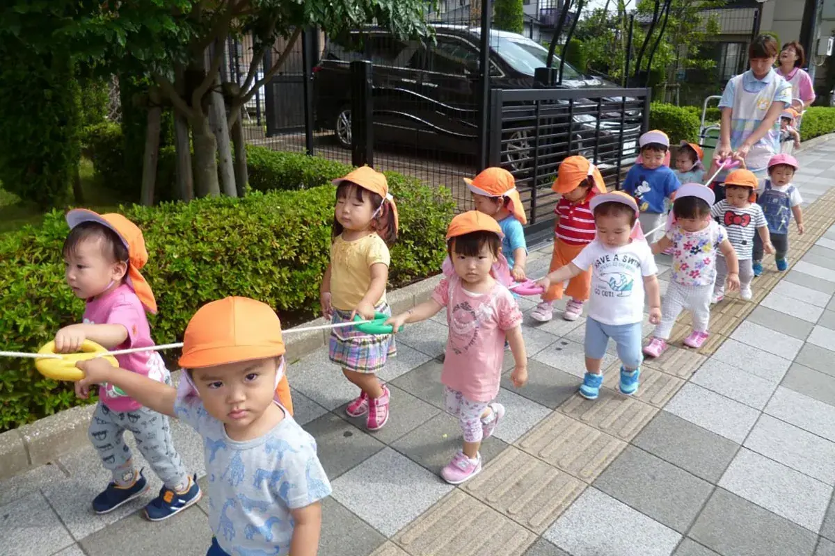 Un gruppo di bambini in Giappone, con cappellini arancioni, cammina in fila tenendo una corda.