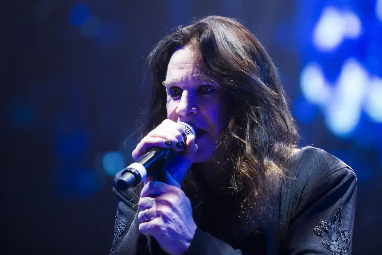 Ozzy Osbourne śpiewa do mikrofonu podczas koncertu, jego długie włosy opadają na twarz.