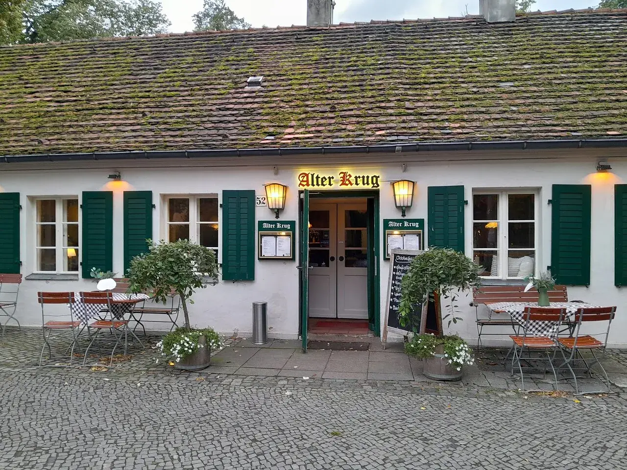 Der "Alter Krug" in Dahlem, ein charmantes Gasthaus mit grünen Fensterläden und einem moosbewachsenen Dach, lädt mit seinen Tischen im Freien zum Verweilen ein.