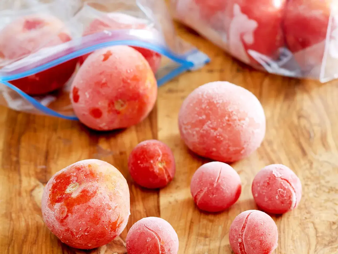 Gefrorene Tomaten liegen auf einem Holzbrett. Einige sind in Gefrierbeuteln, andere einzeln. Perfekt für Soßen und Suppen, wenn man Tomaten einfrieren möchte.