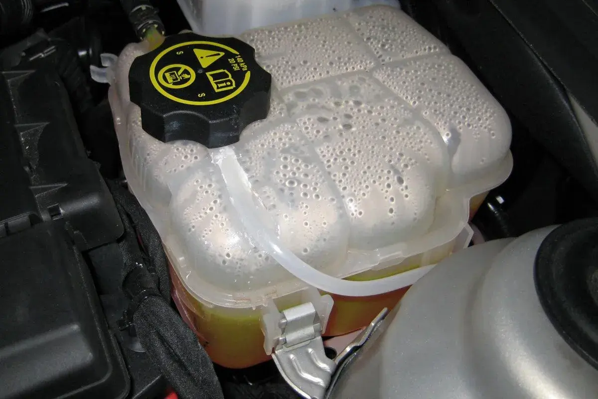 Depósito de expansión del coche con agua y burbujas. El tapón negro tiene símbolos de advertencia.