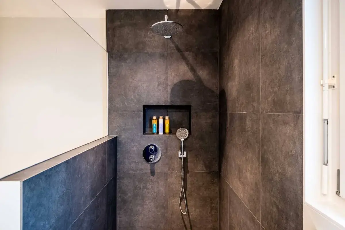 Duchas con murete y cristal. Cabezal de ducha, grifería y nicho con botes de champú. Azulejos oscuros y pared blanca.