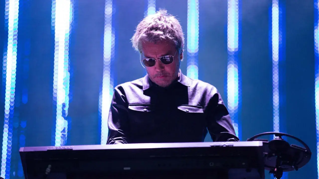 Jean Michel Jarre w Polsce, w okularach przeciwsłonecznych, gra na syntezatorze na tle niebieskich świateł.