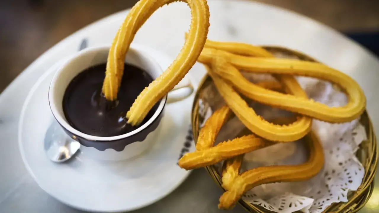 Churros sin gluten dorados y crujientes, uno sumergido en una taza de chocolate caliente espeso.