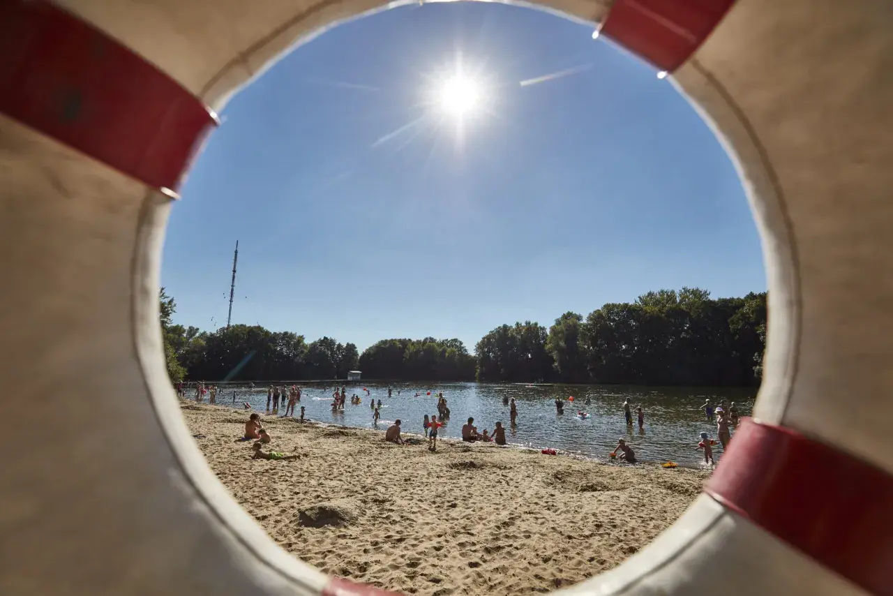 Sonniger Tag am Badesee in Hannover. Menschen planschen im Wasser und entspannen am Sandstrand, gesehen durch einen Rettungsring.