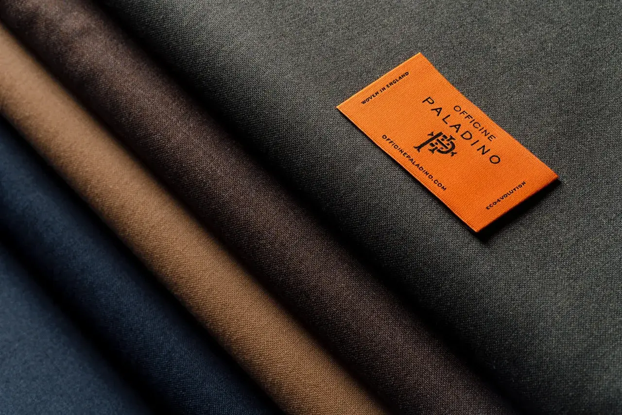 A selection of different types of wool fabric in shades of blue, brown, and grey, with an orange tag from Officine Paladino.