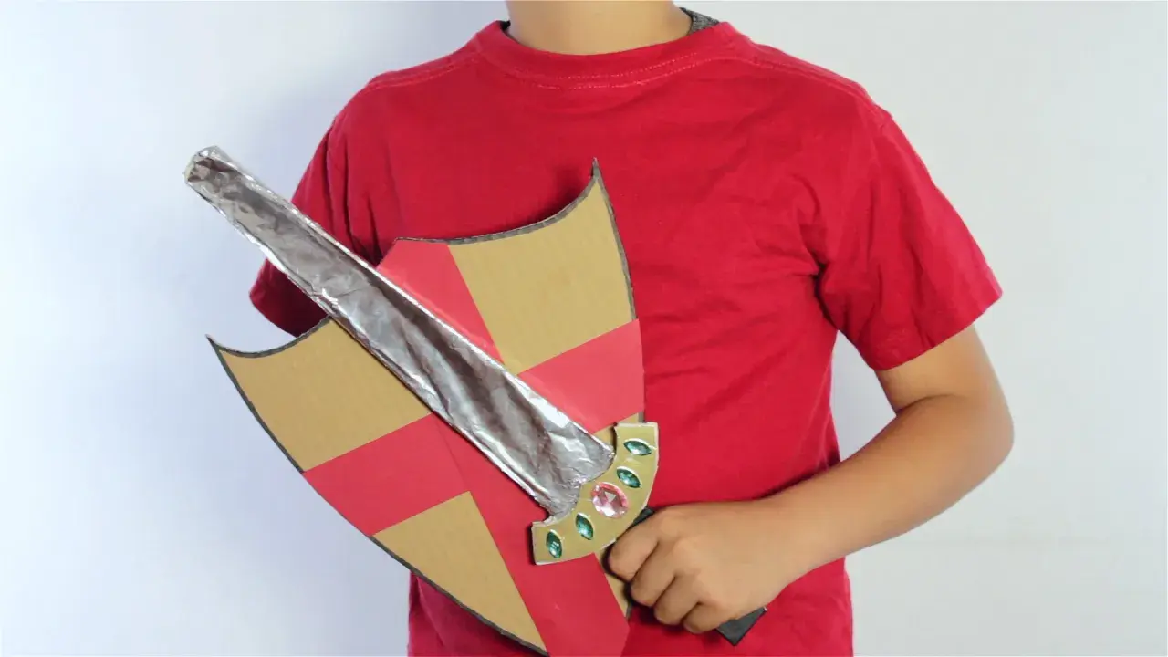 Un niño con camiseta roja sostiene un escudo de cartón con detalles rojos y dorados y una espada de cartón plateada, lista para la aventura.