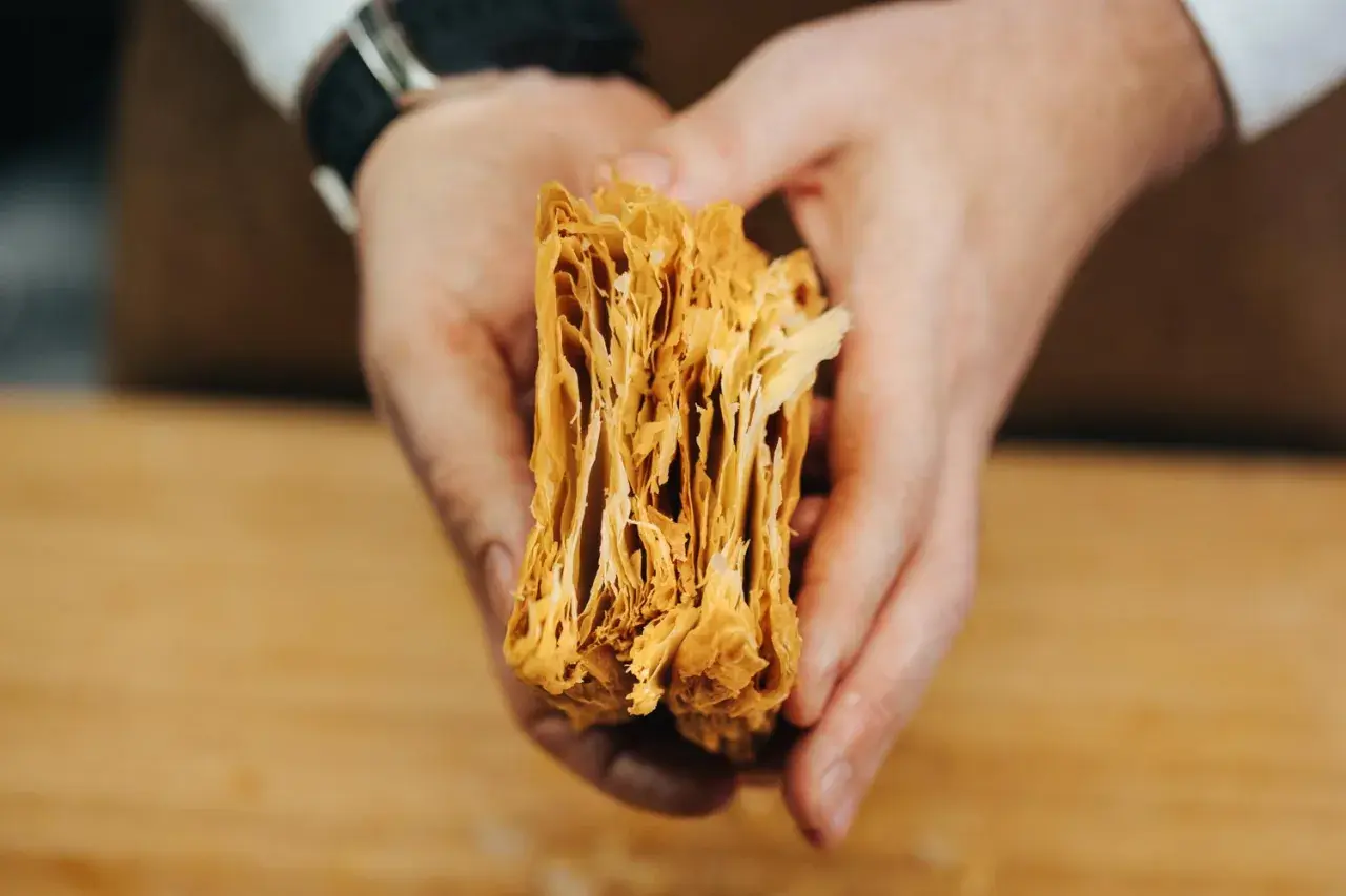 Manos sostienen un trozo de masa de hojaldre casera, mostrando sus finas y crujientes capas doradas.