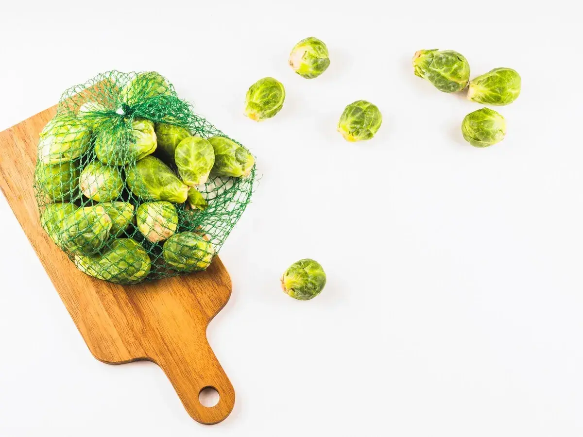 Una bolsa de malla verde llena de coles de Bruselas frescas descansa sobre una tabla de cortar de madera. Algunas coles de Bruselas están esparcidas alrededor.