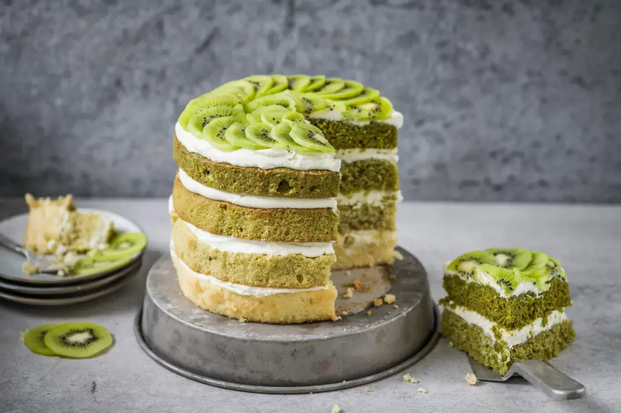 Eine mehrstöckige Matcha Torte mit Kiwi-Scheiben und Sahne. Ein Stück wurde bereits auf einem Teller serviert.