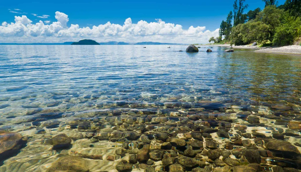 Klares Wasser über Kieselsteinen am Ufer des Lake Taupo. Im Hintergrund eine Insel und bewaldete Hügel unter blauem Himmel mit weißen Wolken.