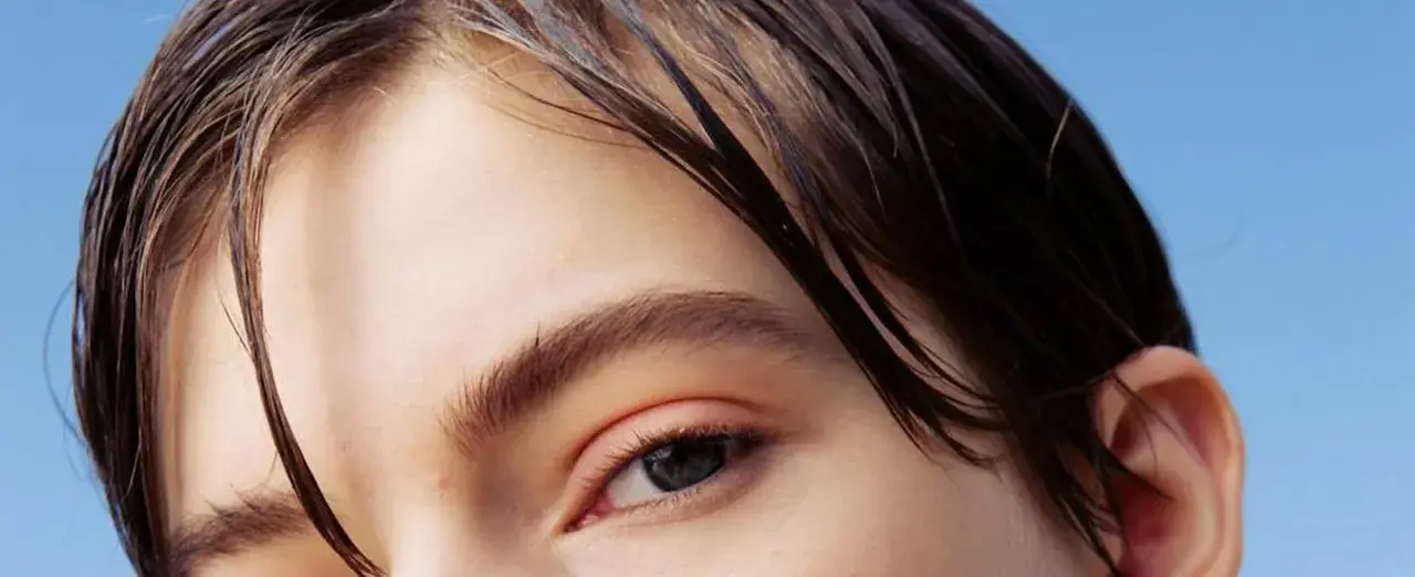 Primer plano de un ojo y cabello oscuro y mojado sobre fondo azul claro.