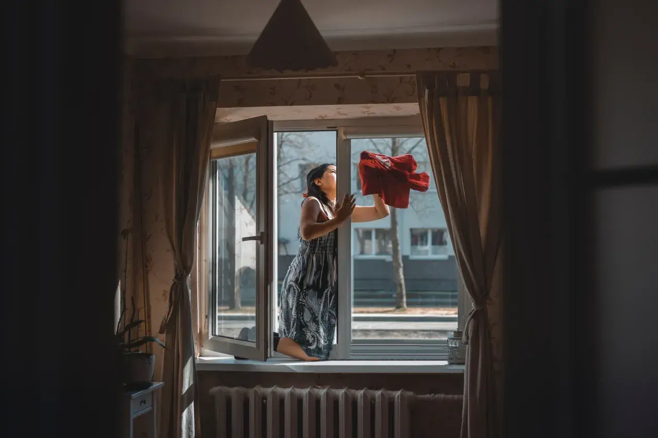 Mujer limpiando una ventana abierta con un paño rojo, parece soñar con barrer.