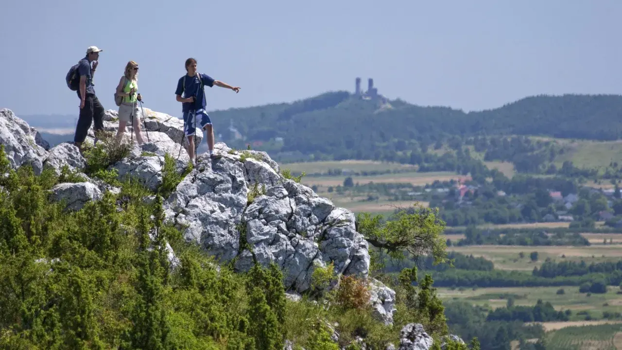 Trzech turystów na skale podziwia krajobraz Gór Świętokrzyskich. Jeden z nich wskazuje kierunek, gdzie w oddali widać ruiny zamku.