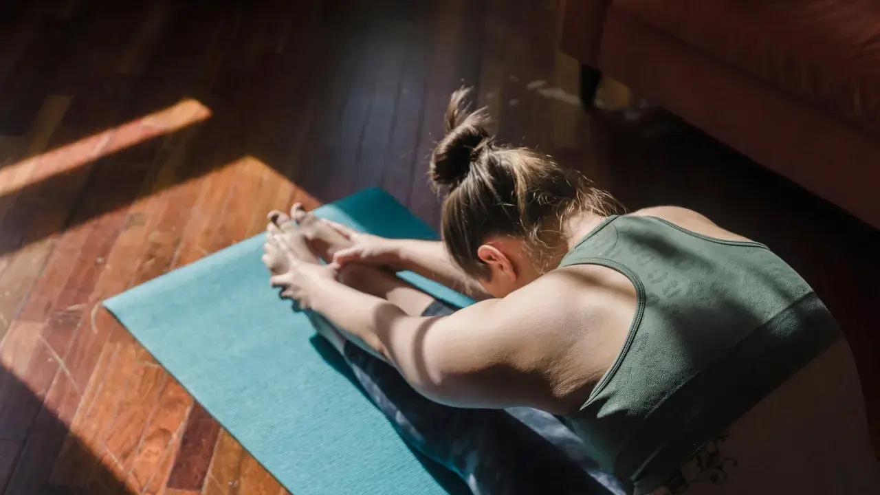 Une femme fait du yoga chez soi, étirant ses jambes sur un tapis bleu, baignée par la lumière du soleil.