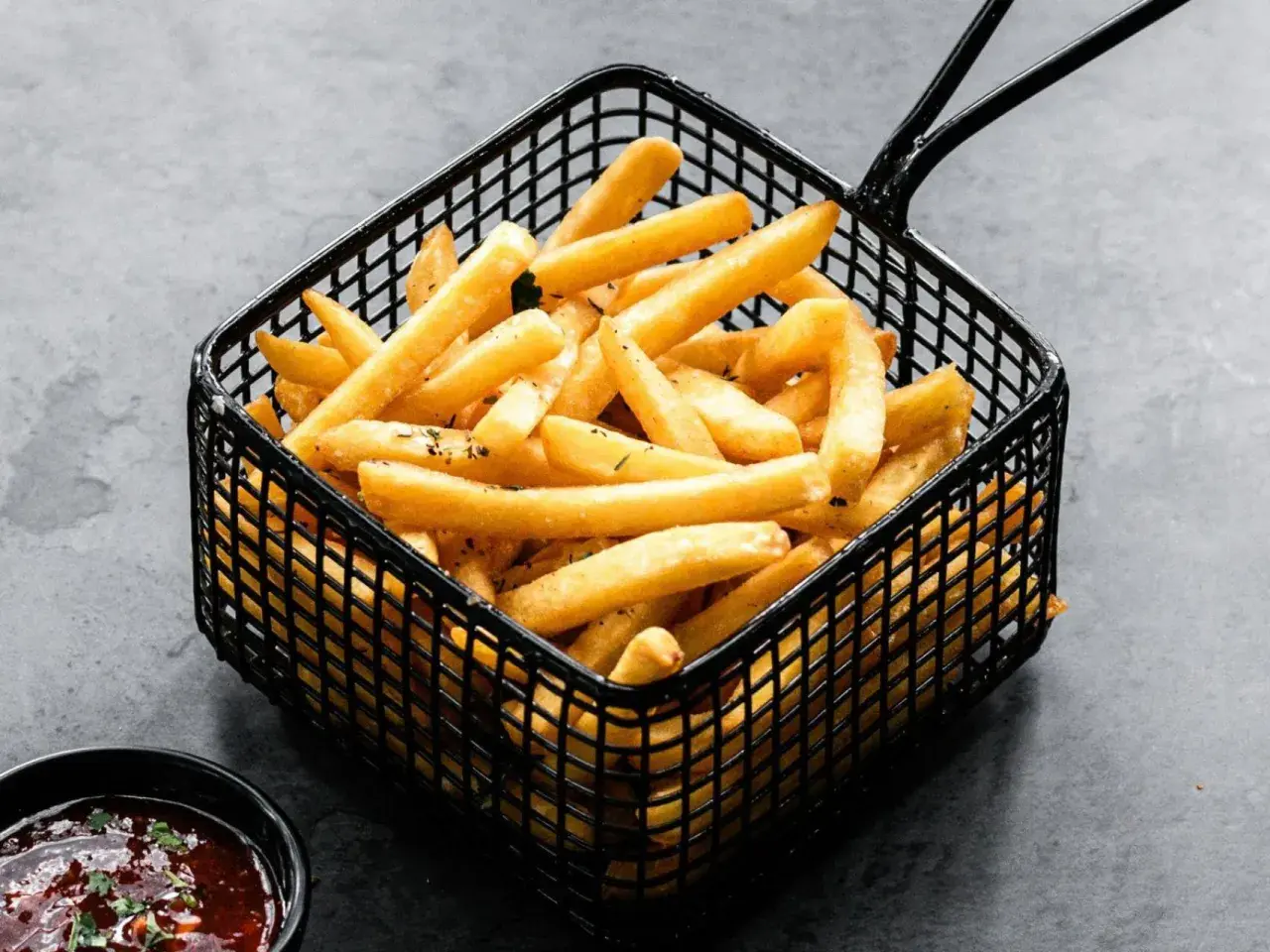 Cesta negra llena de crujientes patatas fritas doradas, con un cuenco de salsa roja al lado.