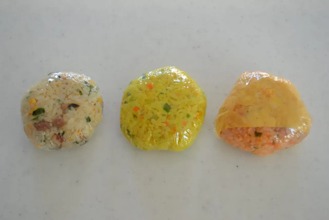 Three plastic-wrapped onigiri sit in a row. The left is white rice with green and brown bits. The middle is yellow rice with orange and green flecks. The right is orange rice with a yellow wrapper. Can you freeze onigiri?