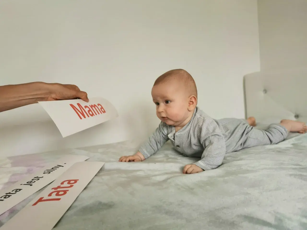 Zaciekawione niemowlę na brzuchu patrzy na kartkę z napisem "Mama". Obok leżą kartki "Tata" i "Tata jest silny".