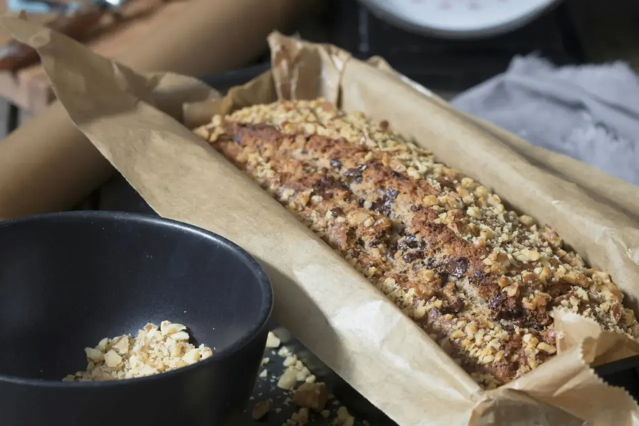 Frisch gebackenes Brot ohne Mehl, bedeckt mit gehackten Nüssen und Schokoladenstückchen, liegt in Backpapier. Daneben eine Schüssel mit Nüssen.