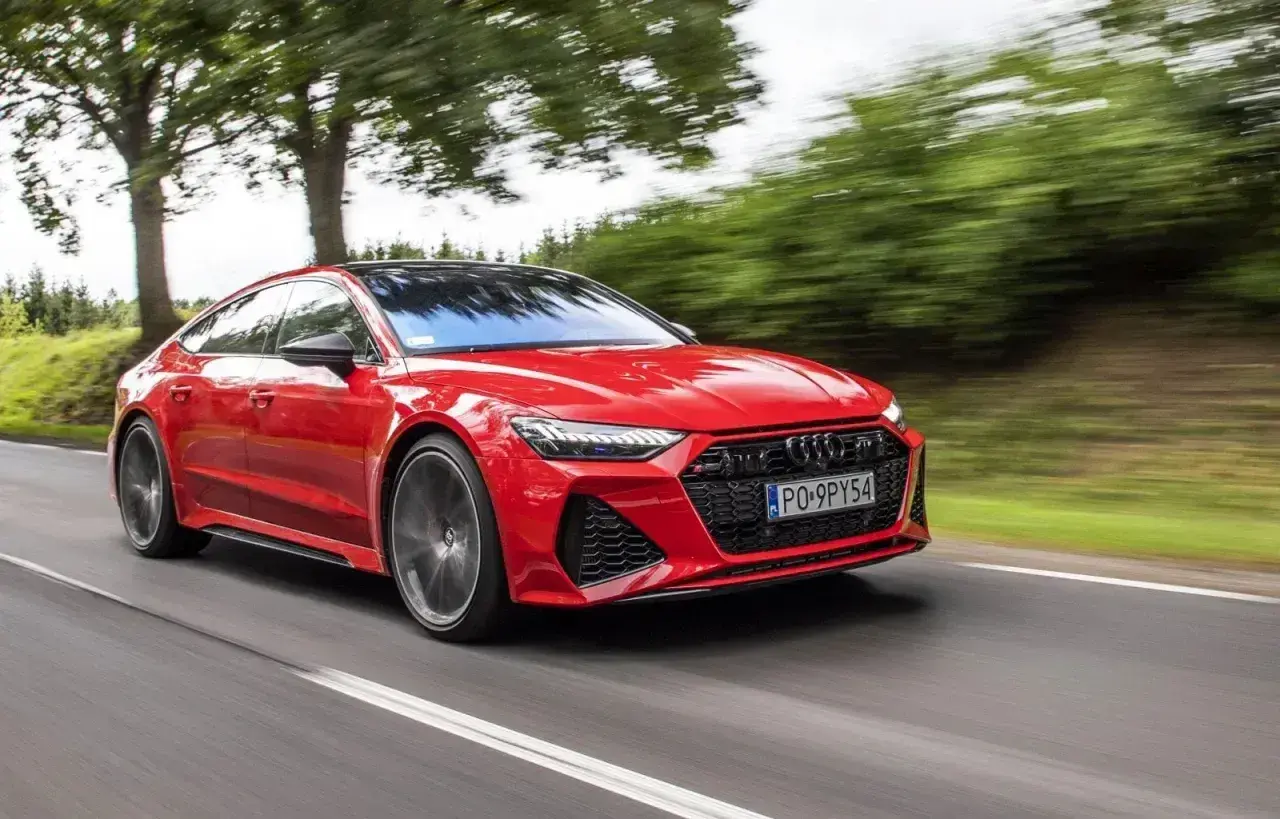 Czerwone Audi RS7 pędzi po drodze, otoczone zielenią drzew.