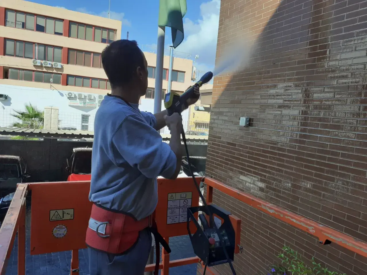 Hombre en plataforma elevadora realiza limpieza de fachadas con agua a presión, dejando la pared de ladrillo brillante y reluciente.