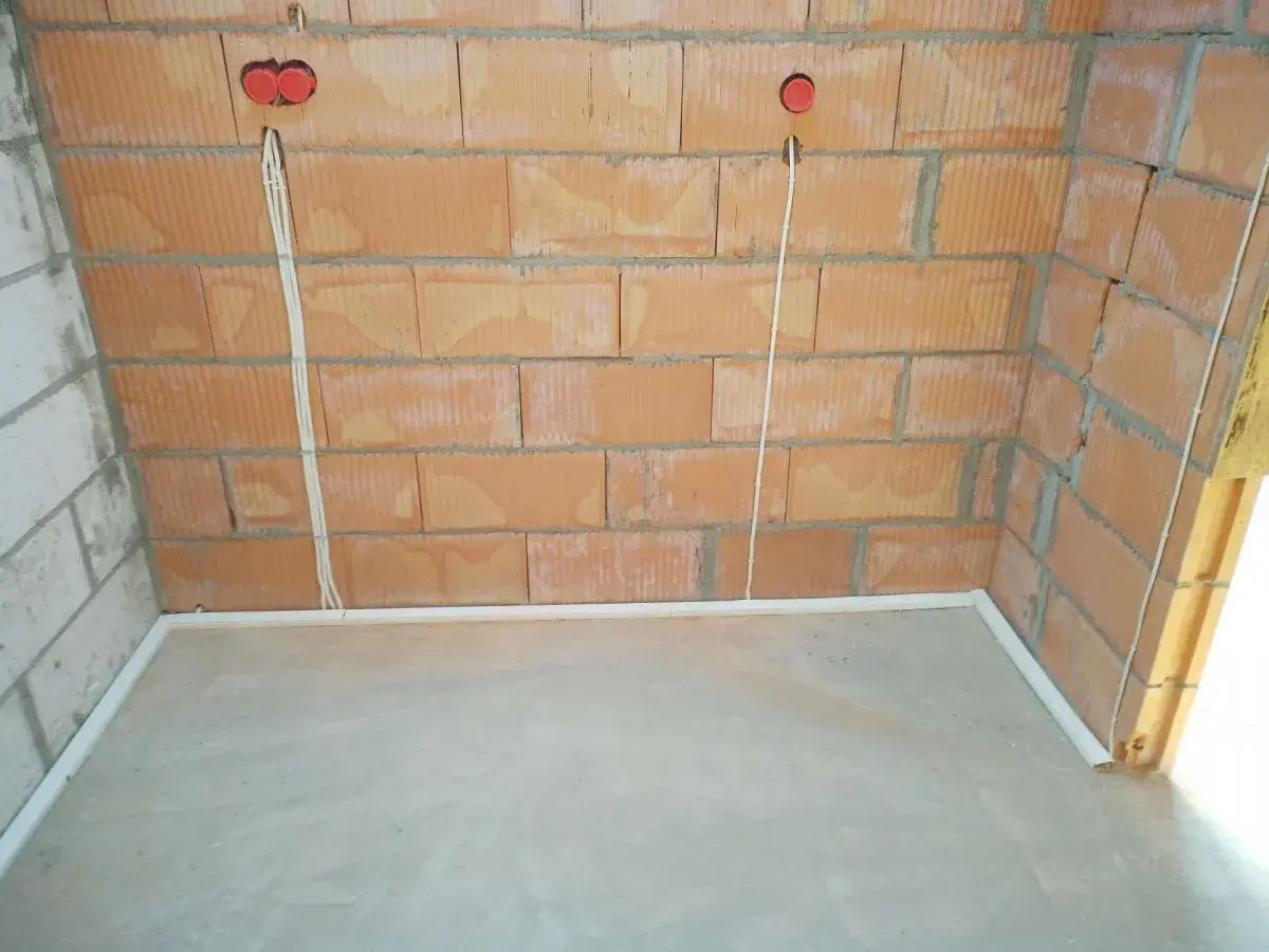 Widoczne przewody w posadzce bez peszla, biegnące od puszek instalacyjnych w ceglanej ścianie do podłogi.