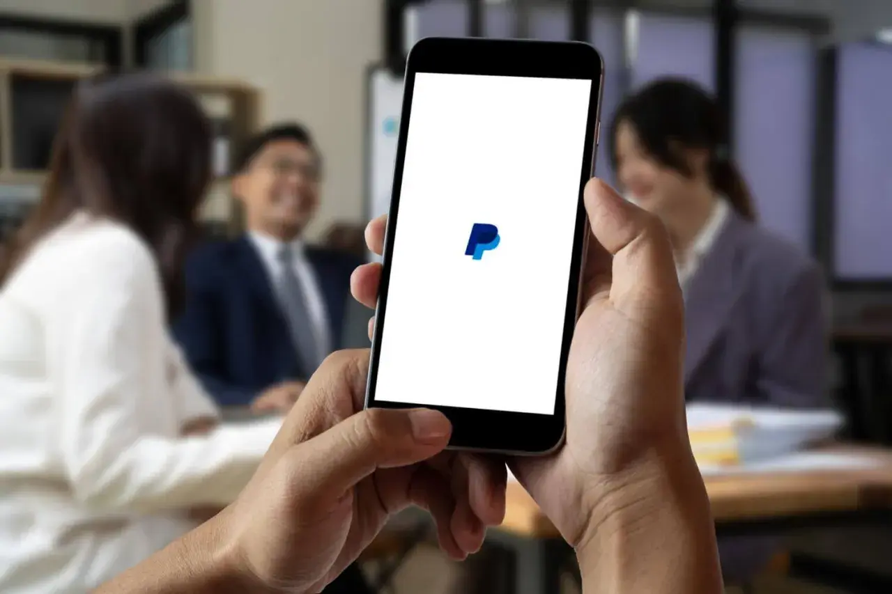 Dłoń trzyma smartfon z logo PayPal na białym ekranie, w tle rozmyte osoby w biurze.