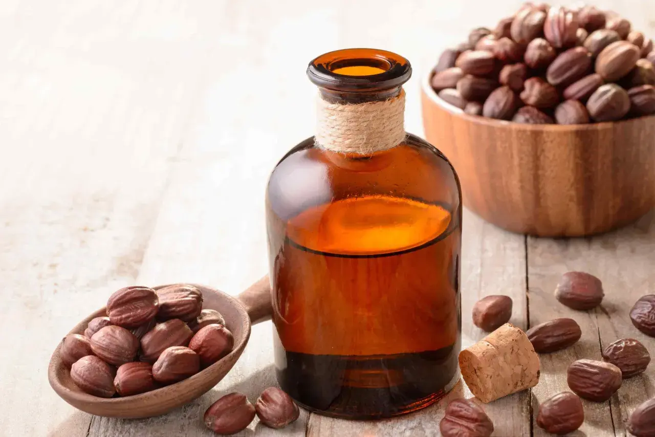 Butelka z bursztynowym płynem, obok drewniana łyżka z nasionami jojoby i miska pełna nasion. Naturalny olej jojoba na włosy.