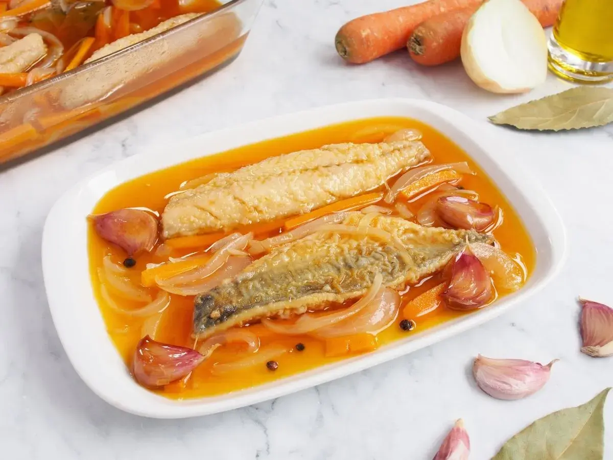 Dos filetes de caballa en escabeche, con zanahoria, cebolla y ajos, en una bandeja blanca.