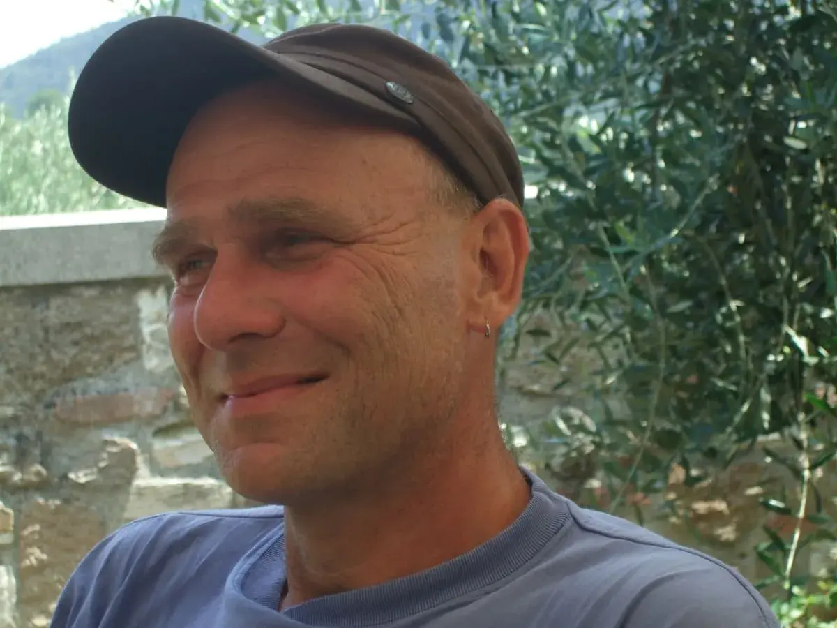 Martin Klein, Autor, lächelt in die Kamera, trägt eine braune Kappe und ein blaues T-Shirt. Im Hintergrund Olivenbäume.