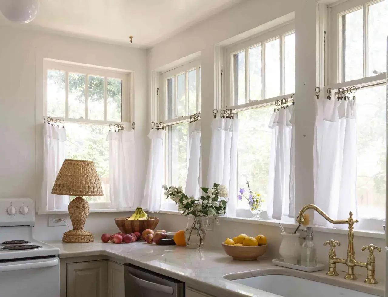 A bright kitchen with white diy cafe curtains framing the windows. A wicker lamp, fruit bowls, and a brass faucet add charm.